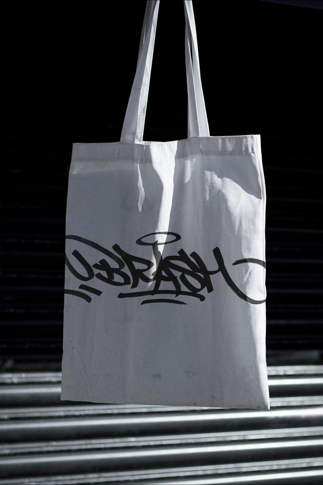 White tote bag mockup with black graffiti style design hanging against dark metal background, ideal for branding presentations.